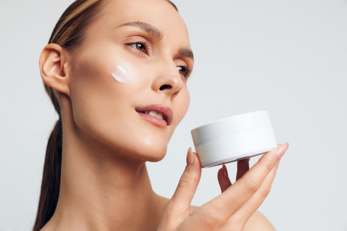 A woman applying cream to her face while holding a white jar, close up